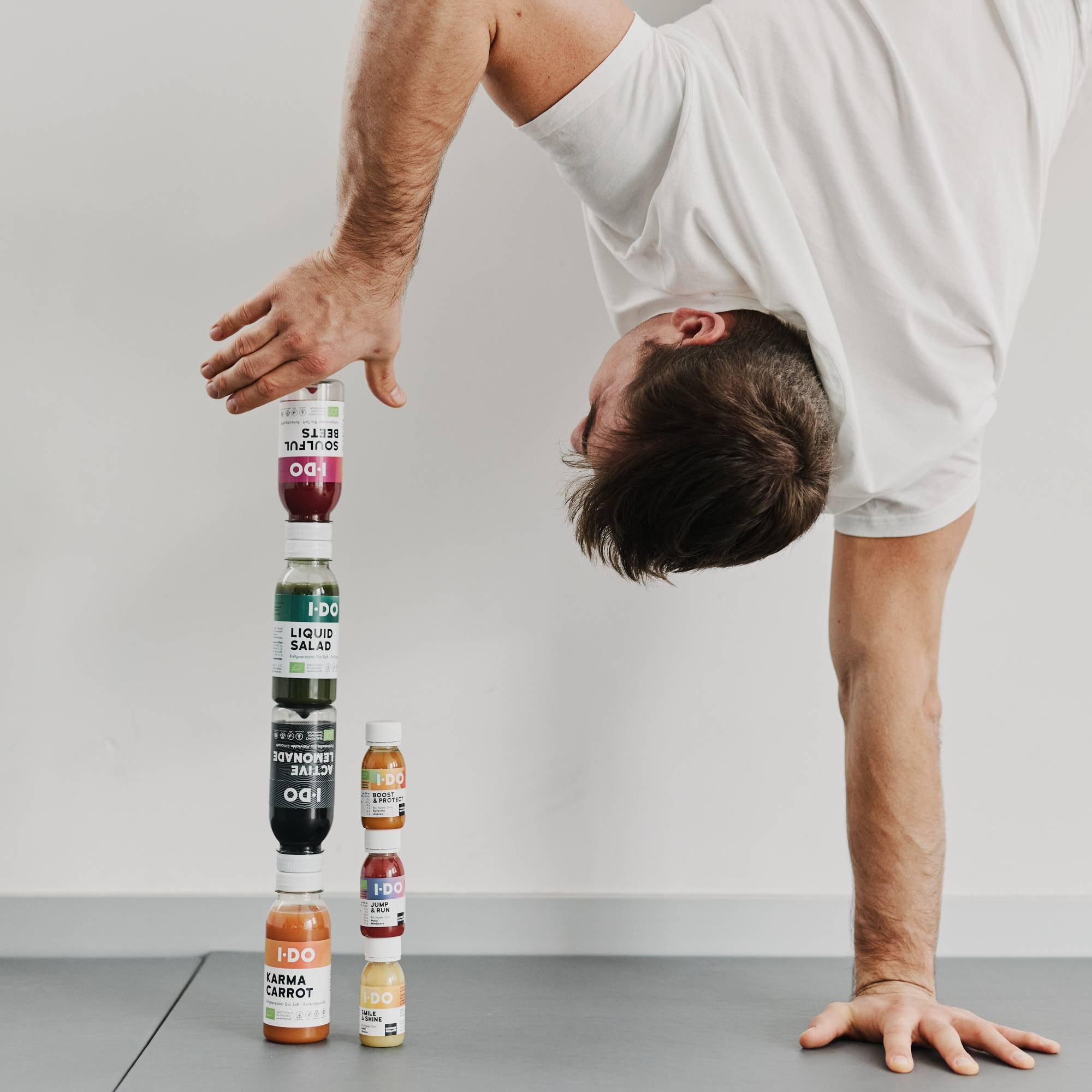 Ein Mann, der einen Handstand macht und Flaschen mit kaltgepresstem Saft stapelt – I·DO